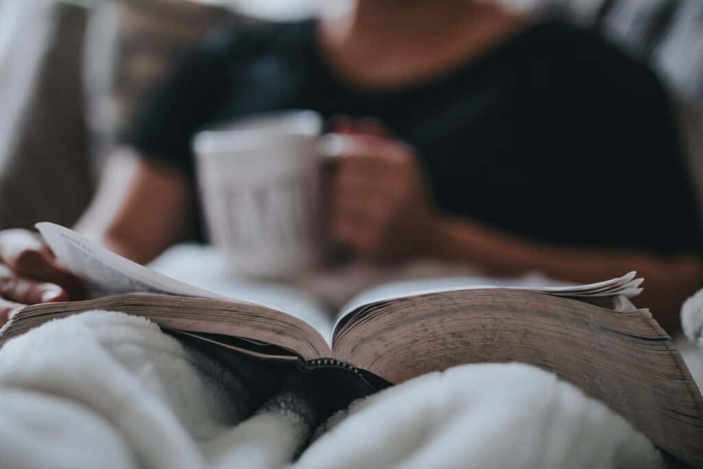 Mies lukee kirjaa sängyssä kahvikuppi toisessa kädessään.